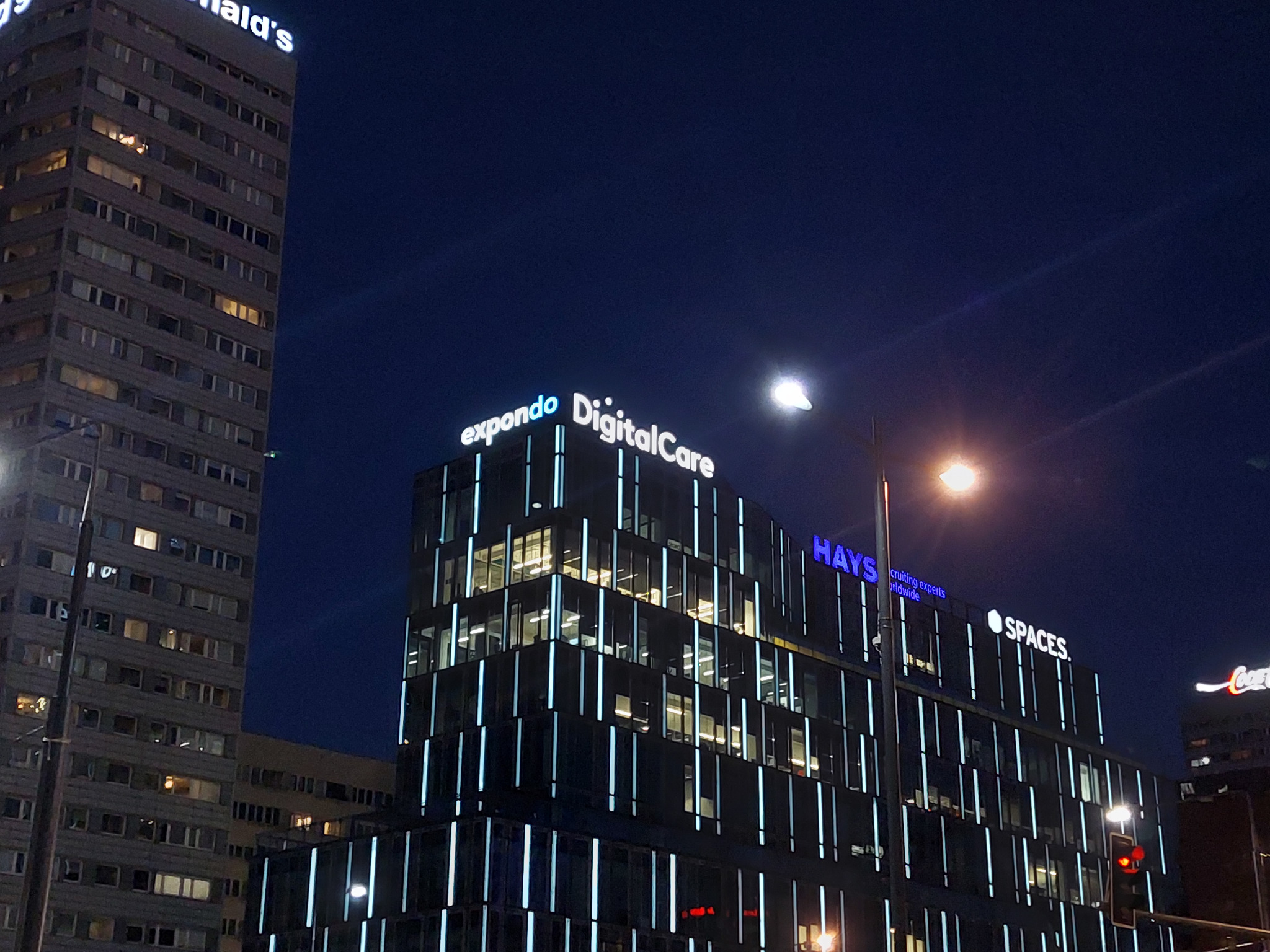 Nocny widok na nowoczesny biurowiec z podświetlonymi pionowymi liniami, na dachu widoczne podświetlane nazwy firm Expando, DigitalCare, Hays i Spaces, w tle starszy budynek z logo McDonald's