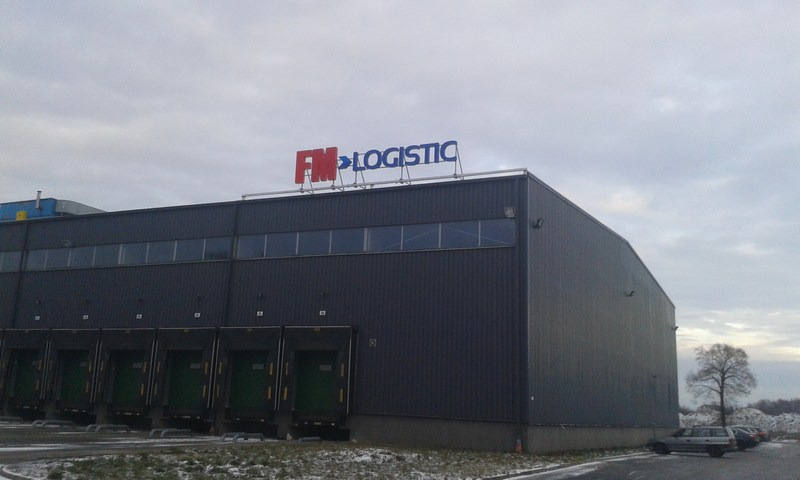 Duży, szary budynek magazynowy z rampami załadunkowymi i logo firmy FM Logistic na dachu, widok z zewnątrz w pochmurny dzień.