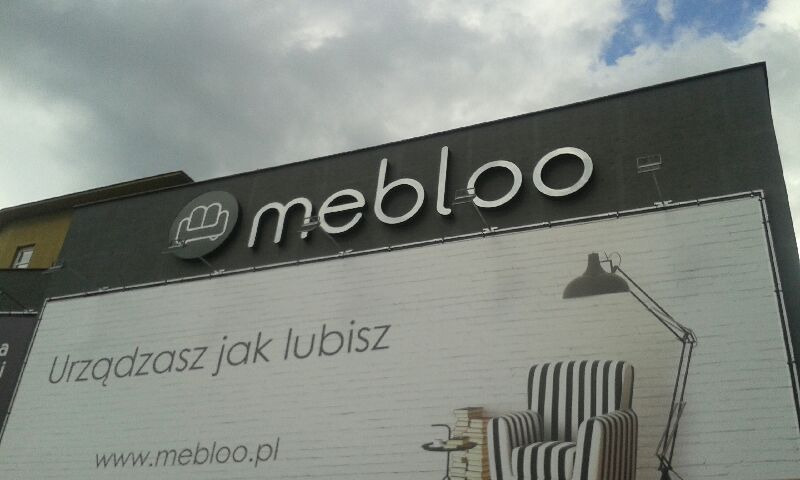 Elewacja budynku z szyldem 'mebloo' i billboardem reklamowym przedstawiającym fotel w paski, lampę podłogową i hasło 'Urządzasz jak lubisz'.