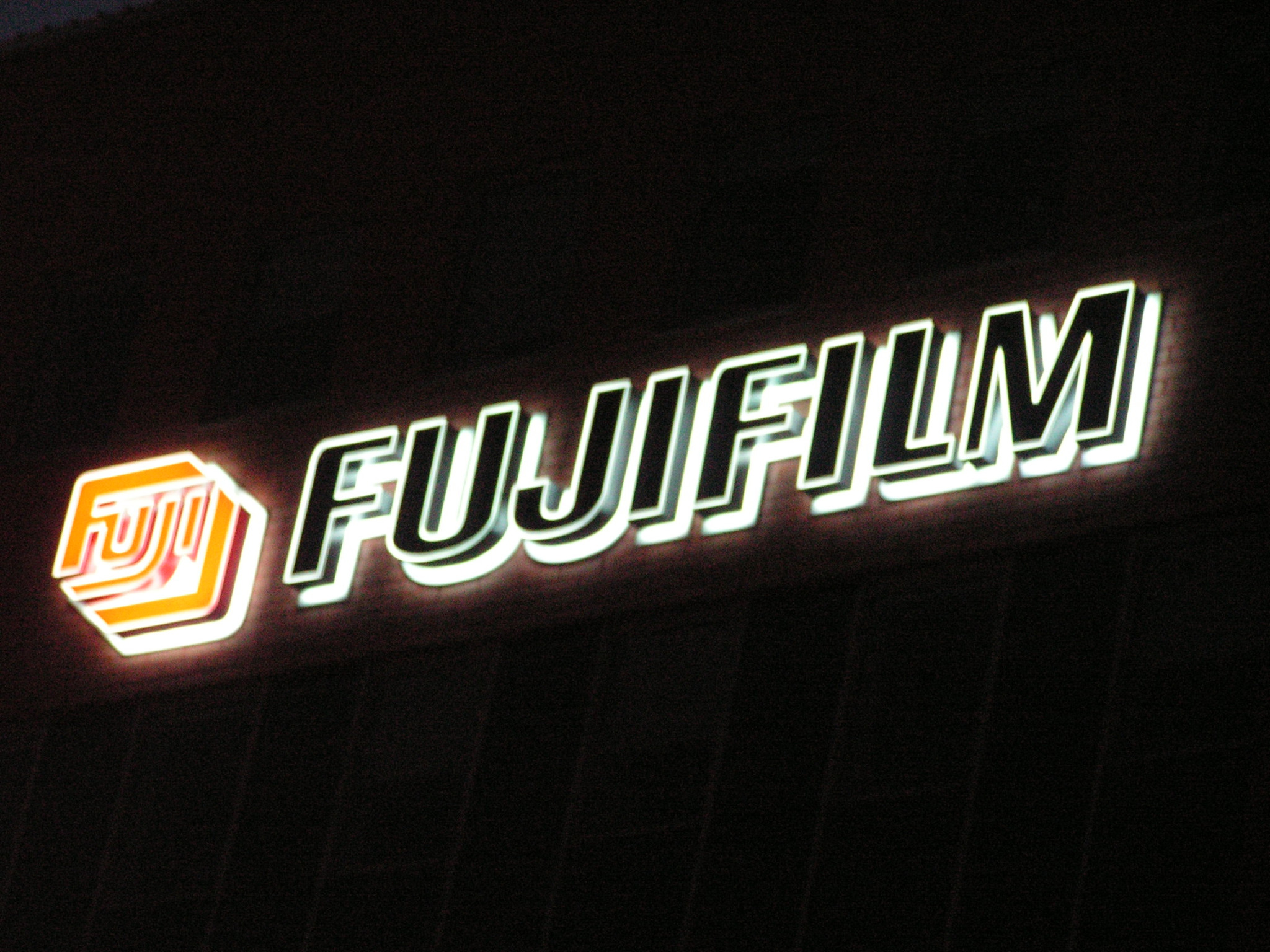 Podświetlany szyld z logo i nazwą Fujifilm na ciemnym tle elewacji budynku, widoczne pionowe prążki na elewacji.
