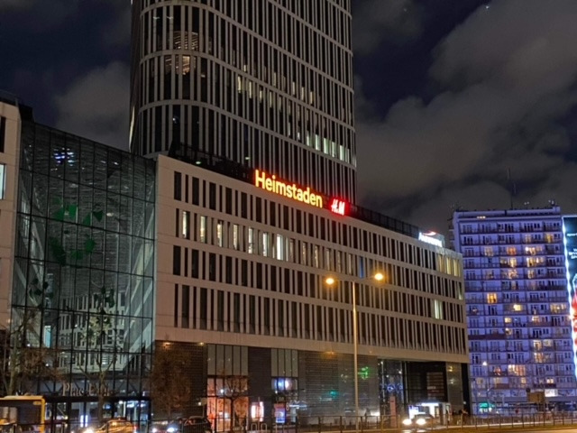 Nocny widok na budynek biurowy z neonowym napisem Heimstaden i logo H&M, otoczony innymi budynkami i rozświetlony ulicznymi lampami.