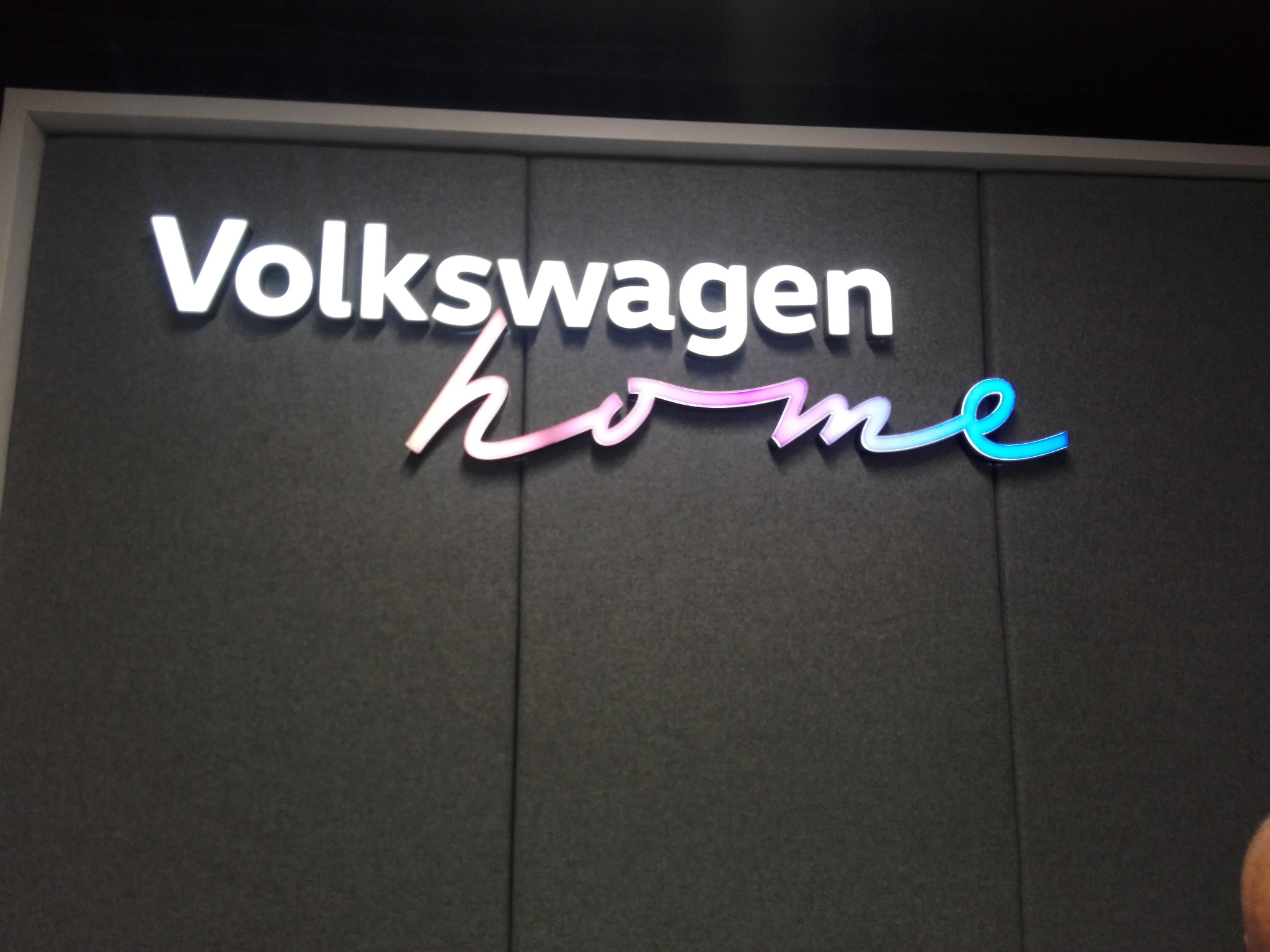 Podświetlany szyld z nazwą Volkswagen Home, litery 'home' w kolorze przechodzącym od różowego do turkusowego, na ciemnoszarym tle.