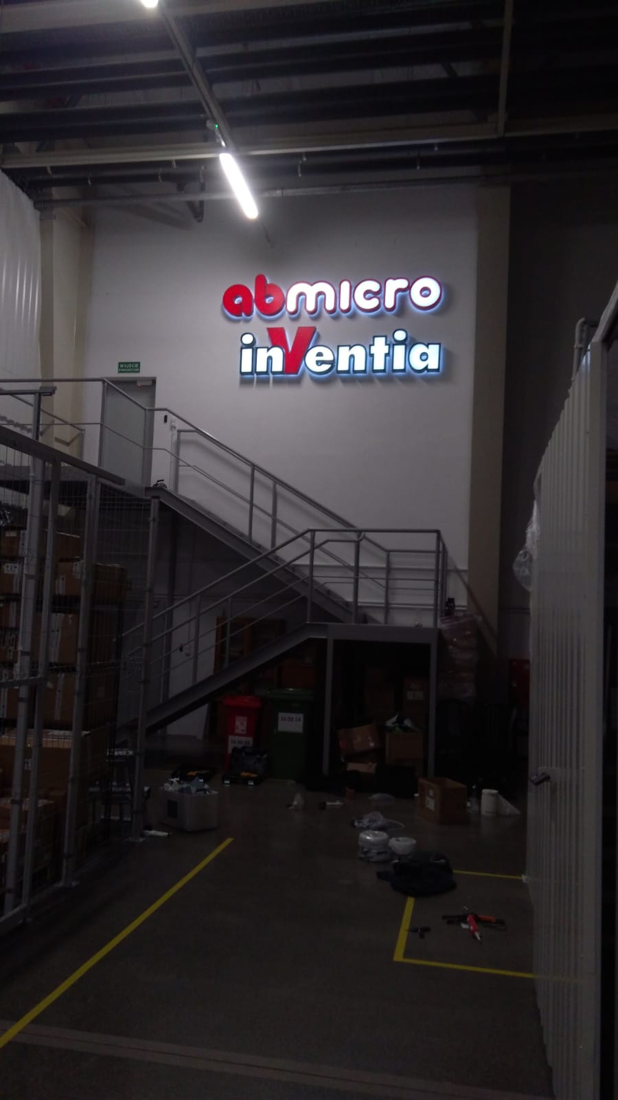 Podświetlany szyld z nazwą firmy 'abmicro inVentia' w hali magazynowej, widoczne schody metalowe i regały z kartonami, oświetlenie sufitowe.
