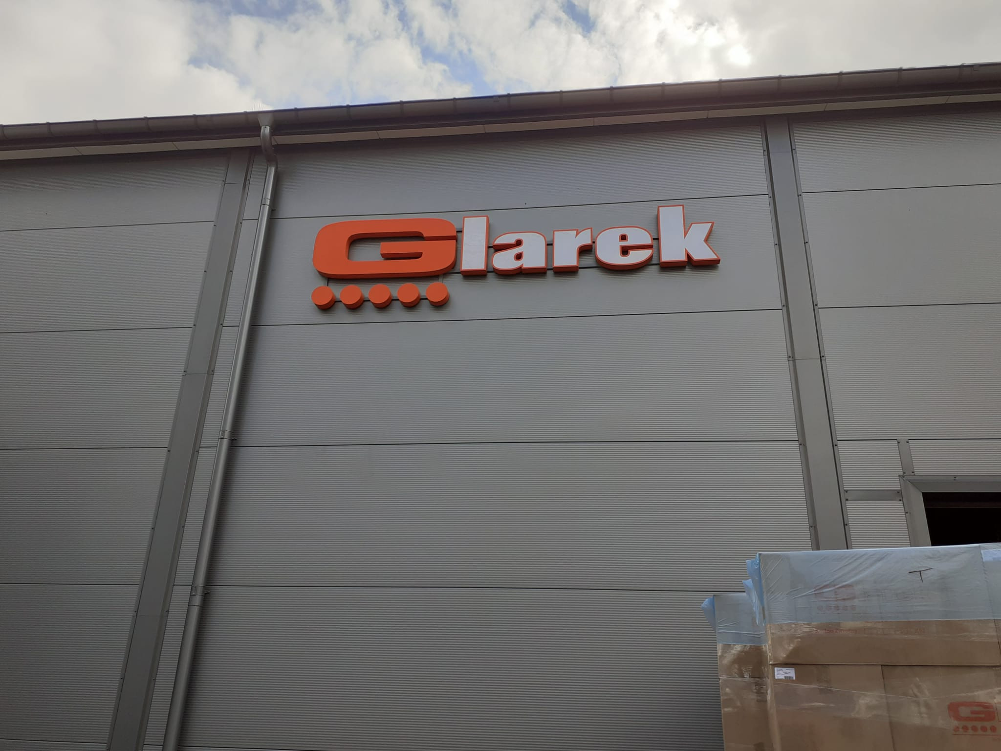 Pomarańczowo-biały szyld z nazwą 'Glarek' umieszczony na szarej, metalowej ścianie budynku przemysłowego pod pochmurnym niebem.