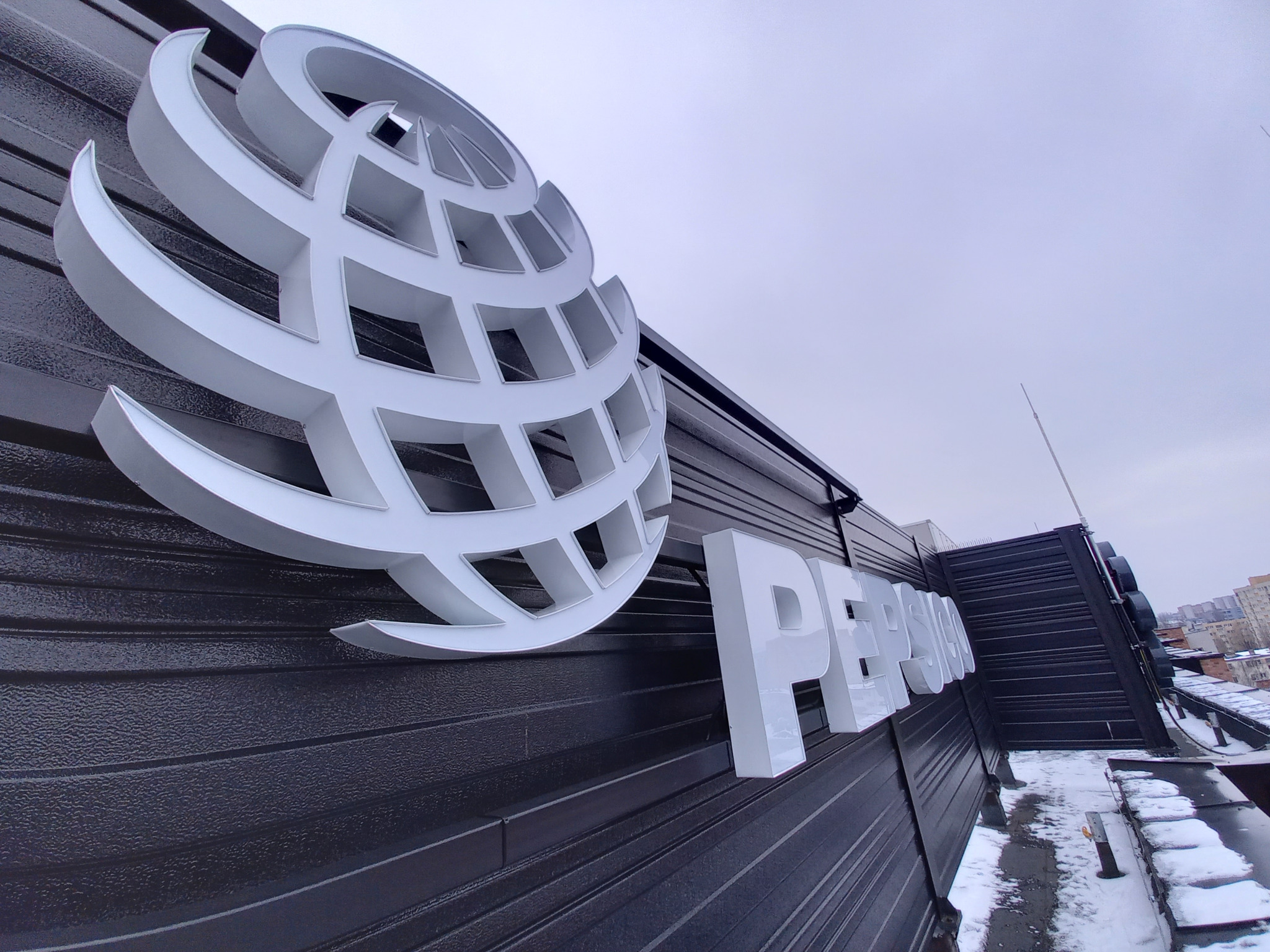 Elewacja budynku z dużym, przestrzennym logo w kształcie globu i napisem w kolorze białym, widoczne z dołu pod kątem, na tle pochmurnego nieba i fragmentów ośnieżonego dachu.