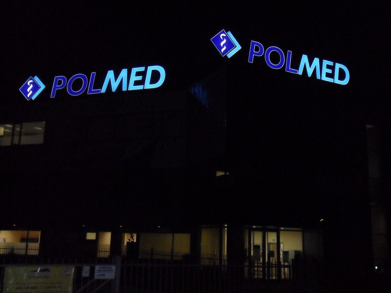 Nocne ujęcie budynku z podświetlanym logo firmy Polmed w kolorze niebieskim, widoczne okna i fragment parkingu.