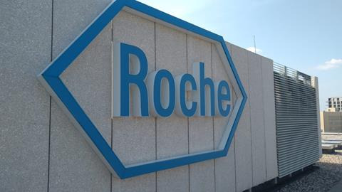 Zewnętrzny szyld firmy Roche z niebieskim logo na szarym tle budynku, widoczne z dołu pod lekkim kątem.
