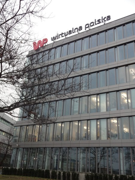 Fasada nowoczesnego biurowca z logo Wirtualnej Polski na dachu, widok z dołu przez gałęzie drzew bez liści.