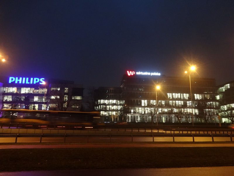 Nocny widok na biurowce z neonowymi szyldami Philips i Wirtualna Polska, z rozmytym autobusem przejeżdżającym na pierwszym planie.