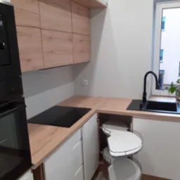 Nowoczesna kuchnia narożna z drewnianymi blatami i białymi frontami szafek, wyposażona w wysuwaną półkę narożną typu 'nerka' oraz czarną płytę indukcyjną i zlewozmywak.