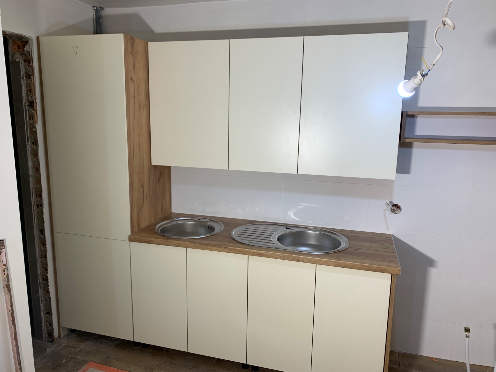 Kuchnia w trakcie montażu: widoczne szafki stojące i wiszące w kolorze kremowym, blat drewnopodobny z dwoma zlewozmywakami, surowe ściany w tle i wisząca żarówka.