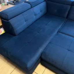 Ciemnoniebieska narożna kanapa z welurowym wykończeniem, widoczna z boku, z elementami dekoracyjnymi na stoliku pomocniczym w tle.