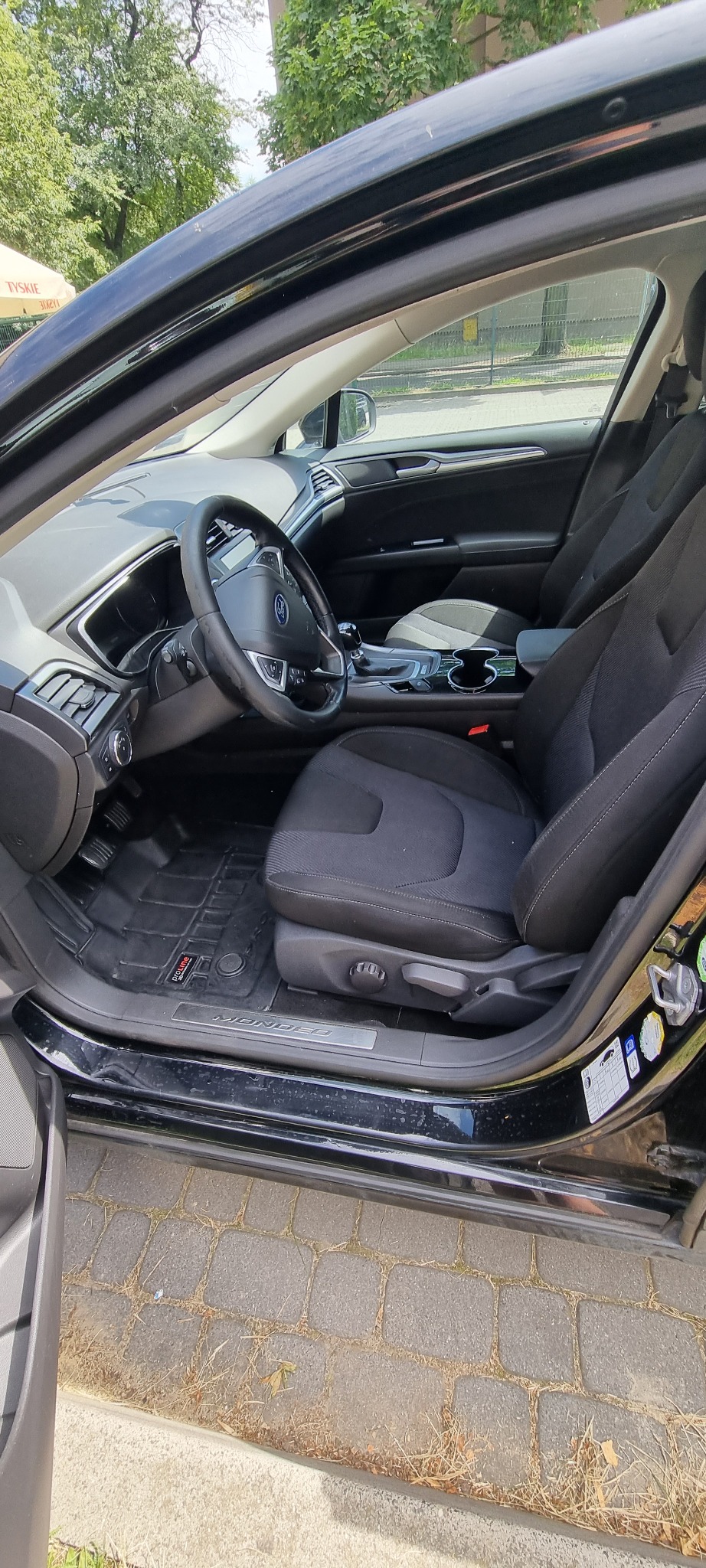 Wnętrze czarnego samochodu Ford Mondeo z widocznymi czarnymi, materiałowymi fotelami, gumowymi dywanikami Proline i otwartymi drzwiami, ukazującymi kostkę brukową na zewnątrz.
