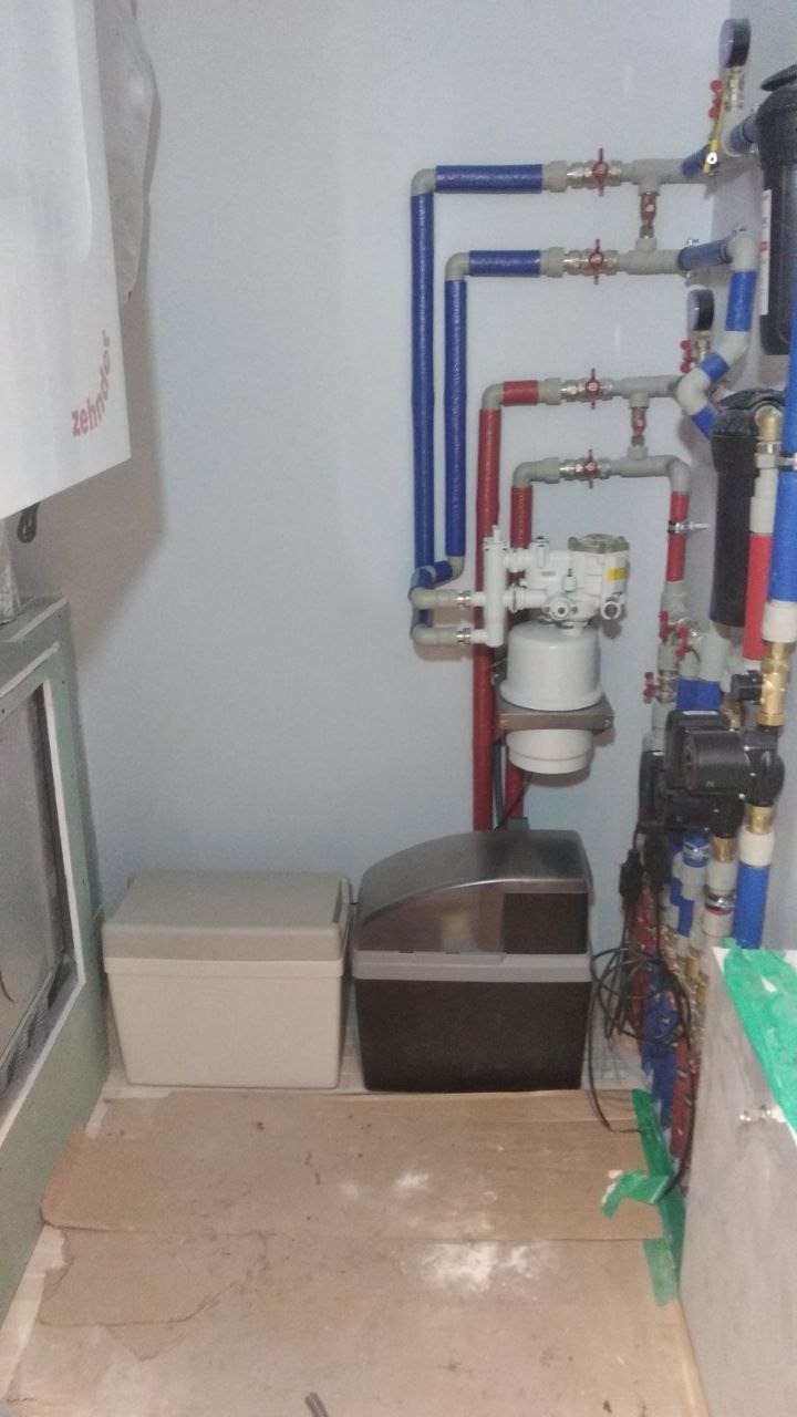 Domowa instalacja z filtrami wody i rurami w kolorach niebieskim i czerwonym, widoczne zbiorniki na podłodze, ściany w kolorze białym.