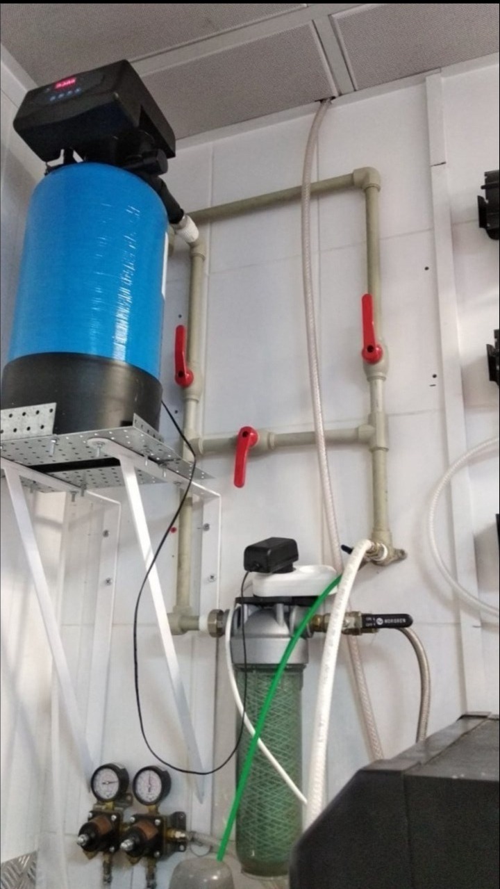 Niebieski zbiornik z czarnym sterownikiem na perforowanej platformie, podłączony do instalacji z rur PCV z czerwonymi zaworami, filtry i manometry widoczne na tle białych płytek.