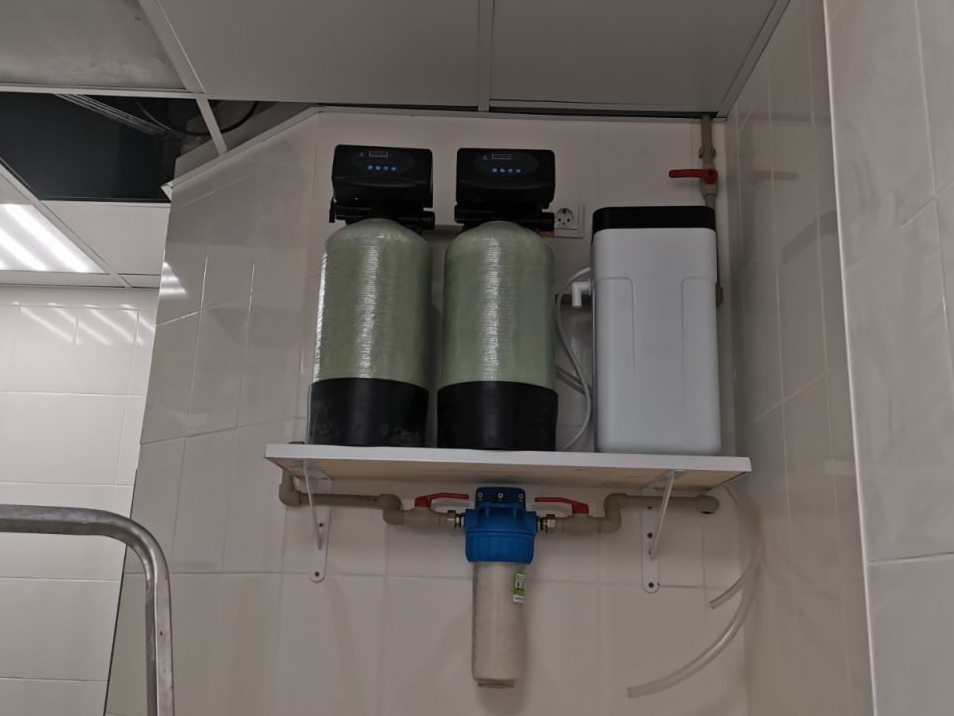 Zestaw filtrów do wody z dwoma cylindrycznymi zbiornikami i białym pojemnikiem na sól, zamontowany na ścianie z białych płytek, widoczny także filtr wstępny pod półką.