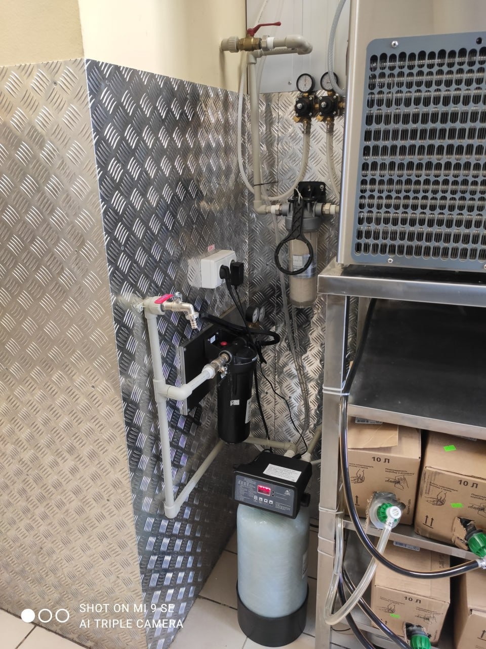 Filtr wody z manometrami i zbiornikiem na tle aluminiowej ściany z ryflowanym wzorem, obok chłodziarka i kartony z napojami.