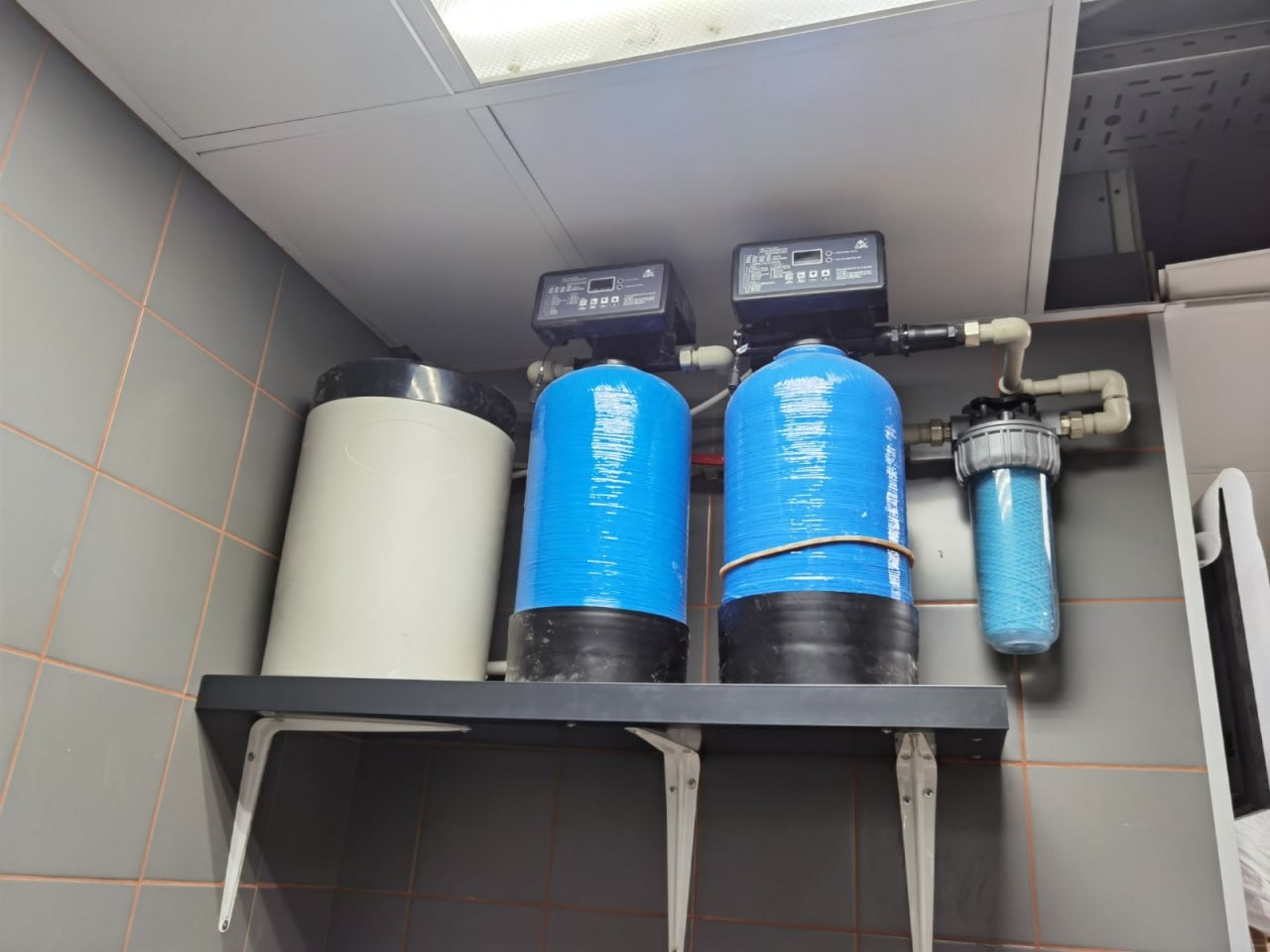 Zestaw filtrów wody z dwoma niebieskimi cylindrycznymi zbiornikami owiniętymi folią, szarym zbiornikiem i przezroczystym filtrem, zamontowany na półce w pomieszczeniu z szarymi płytkami.