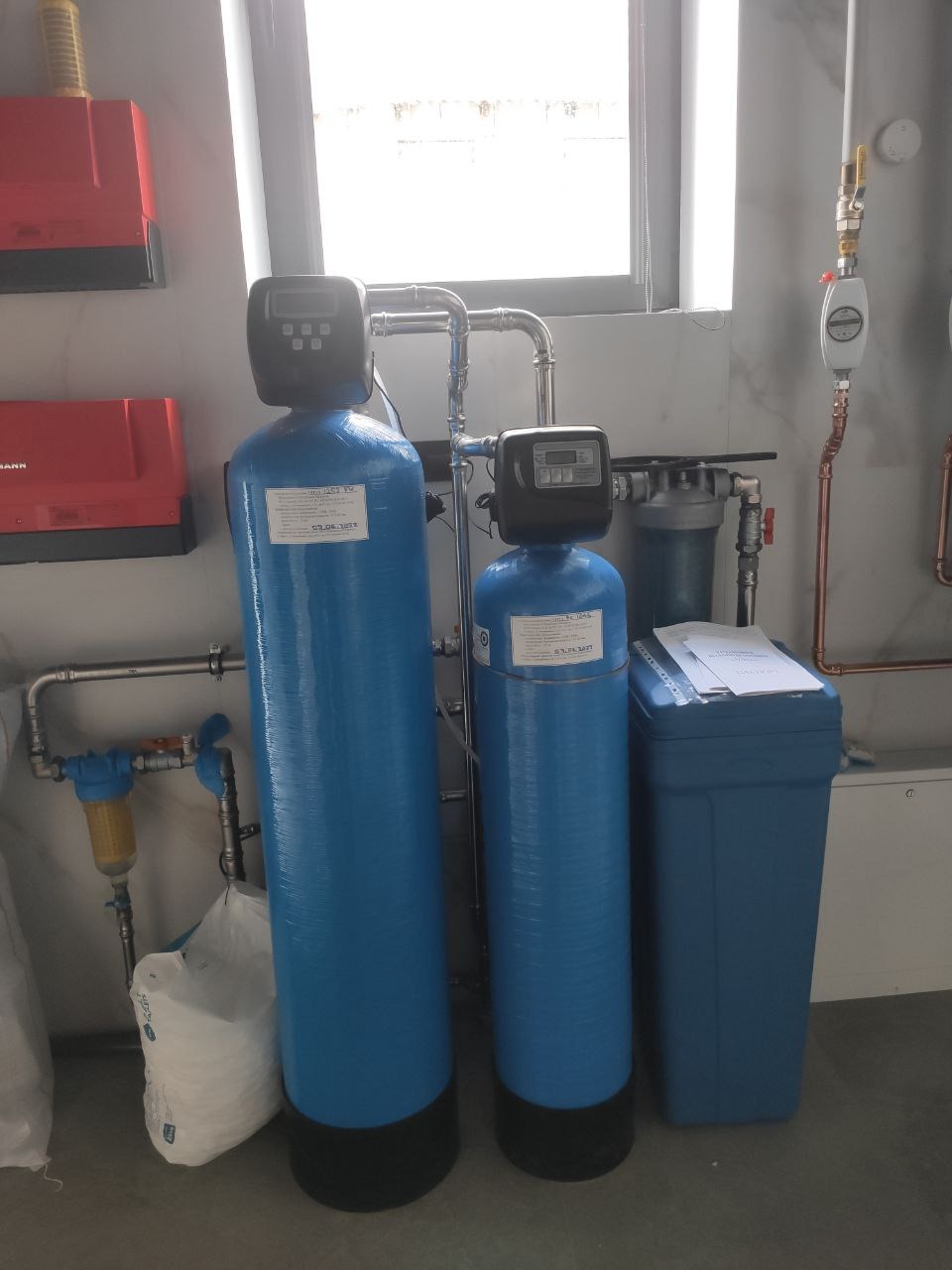 Dwa niebieskie filtry do wody z elektronicznymi panelami sterującymi, połączone rurami, z żółtym filtrem wstępnym i workiem soli obok, na tle białej ściany z oknem i instalacją hydrauliczną.