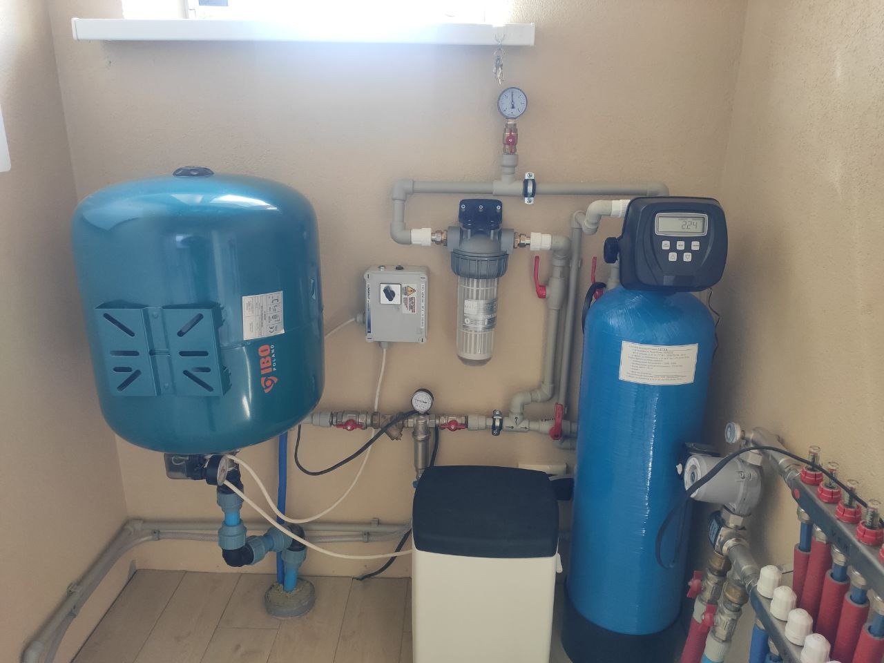 Zestaw urządzeń w kotłowni: hydrofor IBO, filtr narurowy z manometrem, zmiękczacz wody z elektronicznym wyświetlaczem, oraz rozdzielacz ogrzewania podłogowego z rurami PEX.