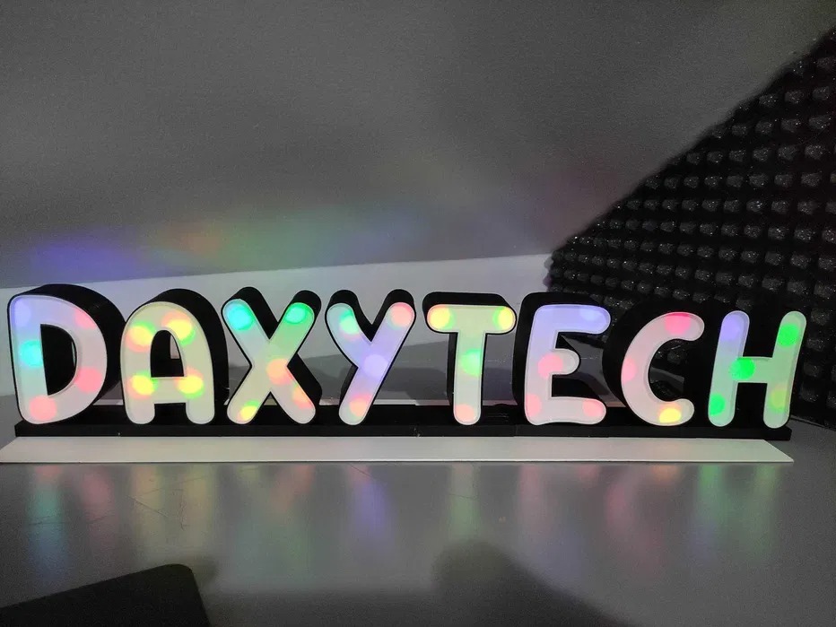 Podświetlany, trójwymiarowy napis 'DAXYTECH' z czarną obudową i białymi, lekko zaokrąglonymi literami wypełnionymi kolorowymi światłami, ustawiony na białej podstawie.