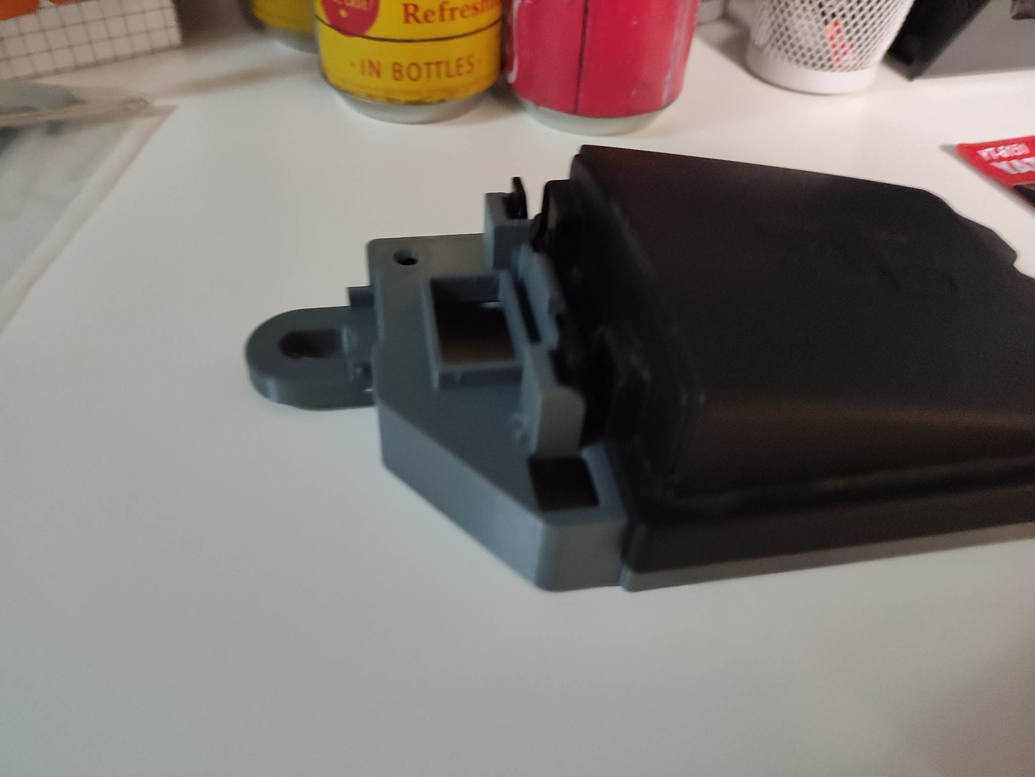 Szary, częściowo wydrukowany element konstrukcyjny z czarnym elementem u góry, widoczne puszki napojów w tle.