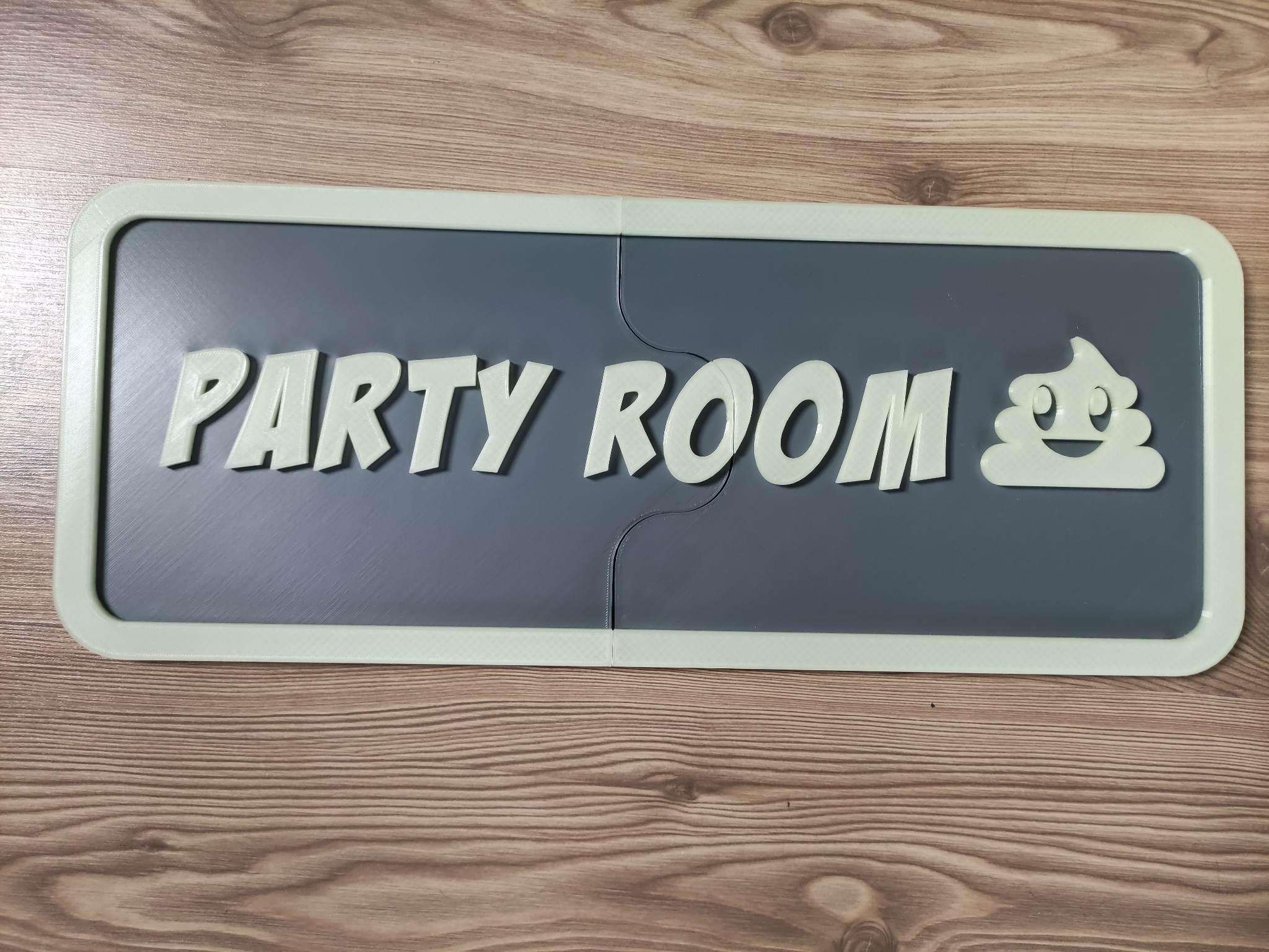 Szary, dwuczęściowy szyld z napisem 'PARTY ROOM' i emoji w kształcie kupy z uśmiechniętą buzią, wykonany prawdopodobnie w Częstochowie przy użyciu technologii druku 3D, leży na drewnianej powierzchni.