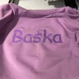 Jasnofioletowa bluza z wyhaftowanym imieniem 'Baśka' w kolorze fioletowym, widoczna pod igłami maszyny do szycia.
