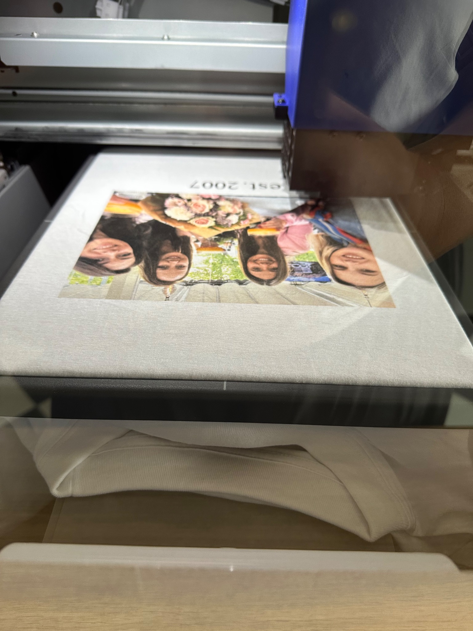 Drukarka UV nanosi kolorowy nadruk ze zdjęciem czterech uśmiechniętych dziewcząt na jasną koszulkę. Widok z góry na proces drukowania.