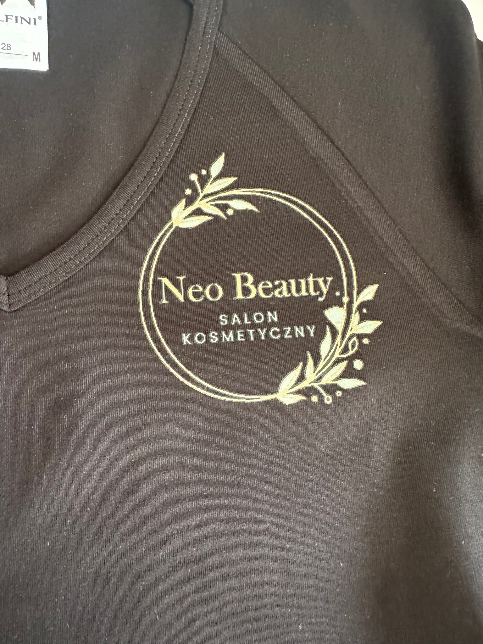 Czarna koszulka z nadrukiem w kolorze złota: 'Neo Beauty. Salon Kosmetyczny', otoczona wiankiem z liści. Widoczny fragment metki.