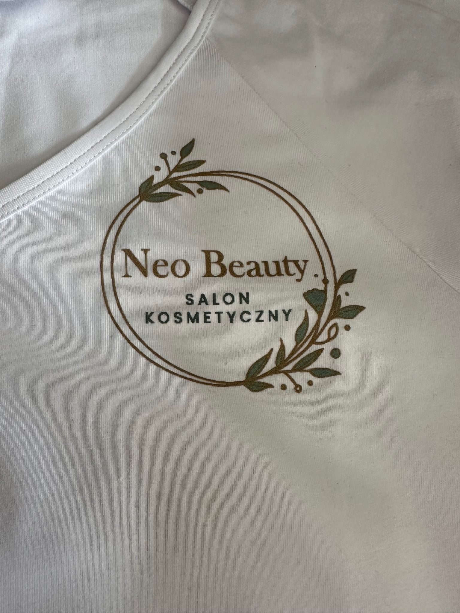 Biała koszulka z nadrukiem logo 'Neo Beauty Salon Kosmetyczny' w okrągłej ramce z motywem roślinnym, wykonanym w odcieniach brązu i zieleni.