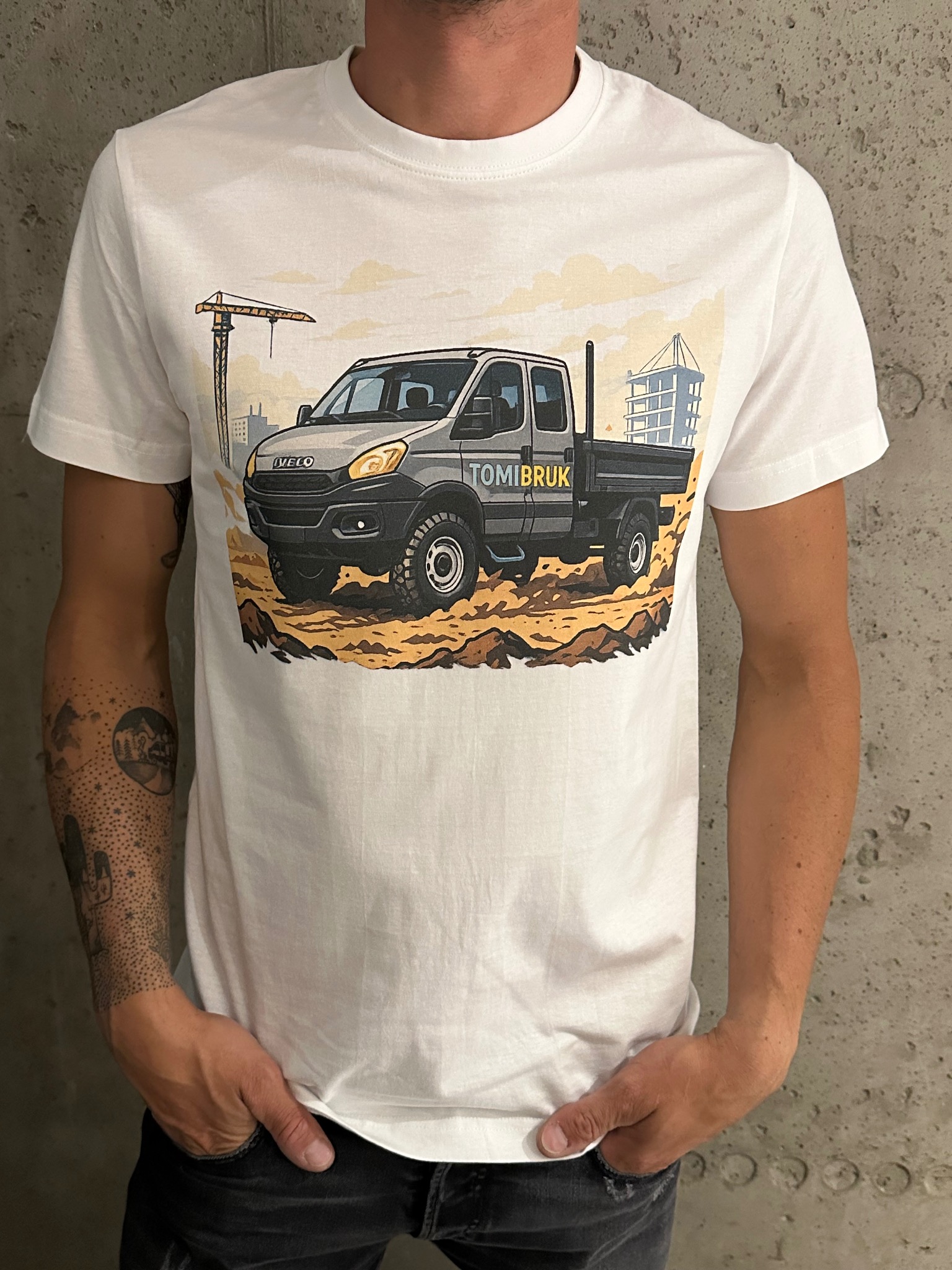 Biała koszulka z nadrukiem przedstawiającym szary samochód ciężarowy na tle krajobrazu miejskiego, noszona przez mężczyznę z tatuażami na ramieniu.