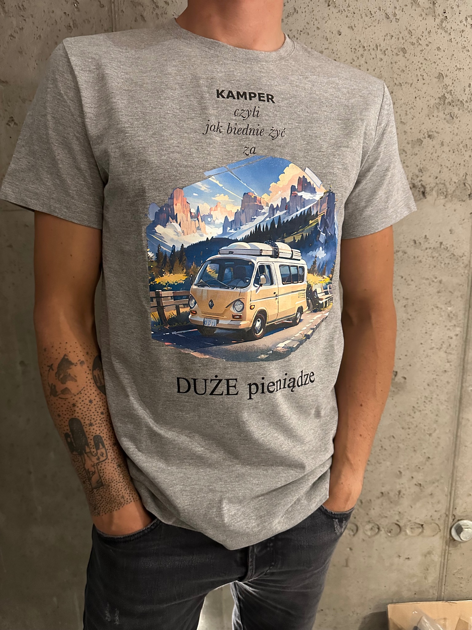 Szara koszulka z nadrukiem retro kampera na tle gór, z hasłem 'Kamper czyli jak biednie żyć za duże pieniądze'. Mężczyzna z tatuażem na ramieniu prezentuje produkt.