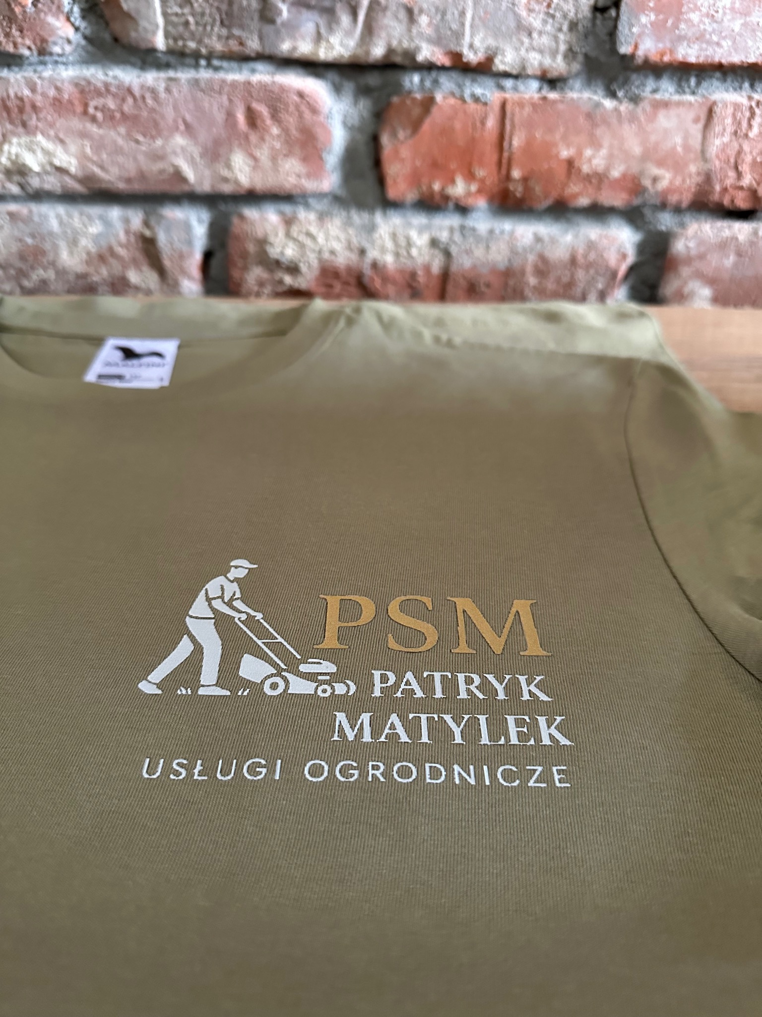 Khaki koszulka z białym nadrukiem: sylwetka ogrodnika z kosiarką, napis 'PSM Patryk Matylek Usługi Ogrodnicze'. Tło: ceglana ściana.