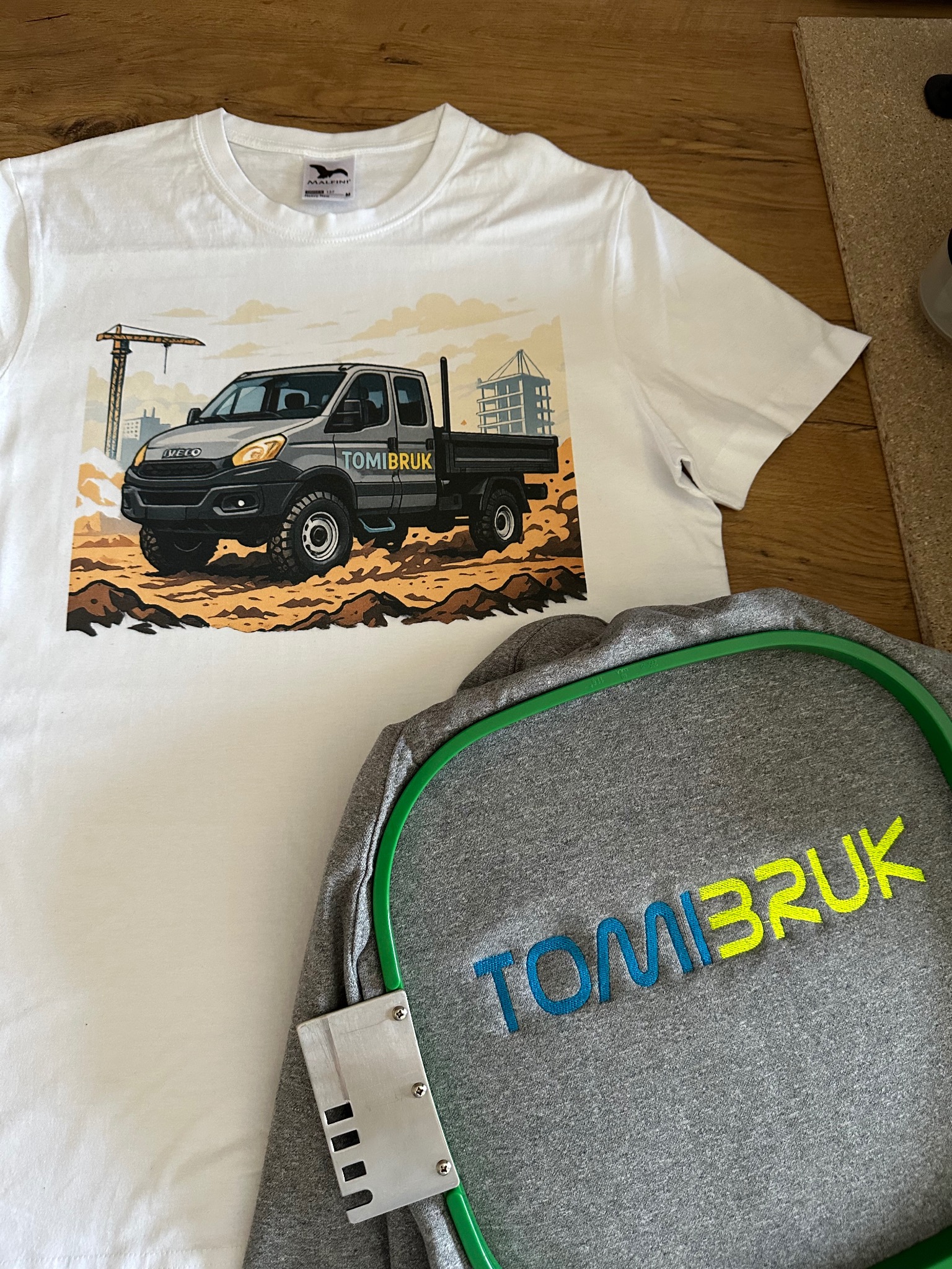 Biała koszulka z nadrukiem ciężarówki Iveco i szara bluza z haftowanym napisem 'TOMIBRUK' na drewnianym tle.