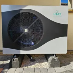 Zewnętrzna jednostka pompy ciepła, model HAPE Ciepły Dom, zainstalowana na betonowych blokach na brukowanym podłożu z drobnym żwirem, widoczny fragment elewacji budynku i zwinięty wąż ogrodowy.