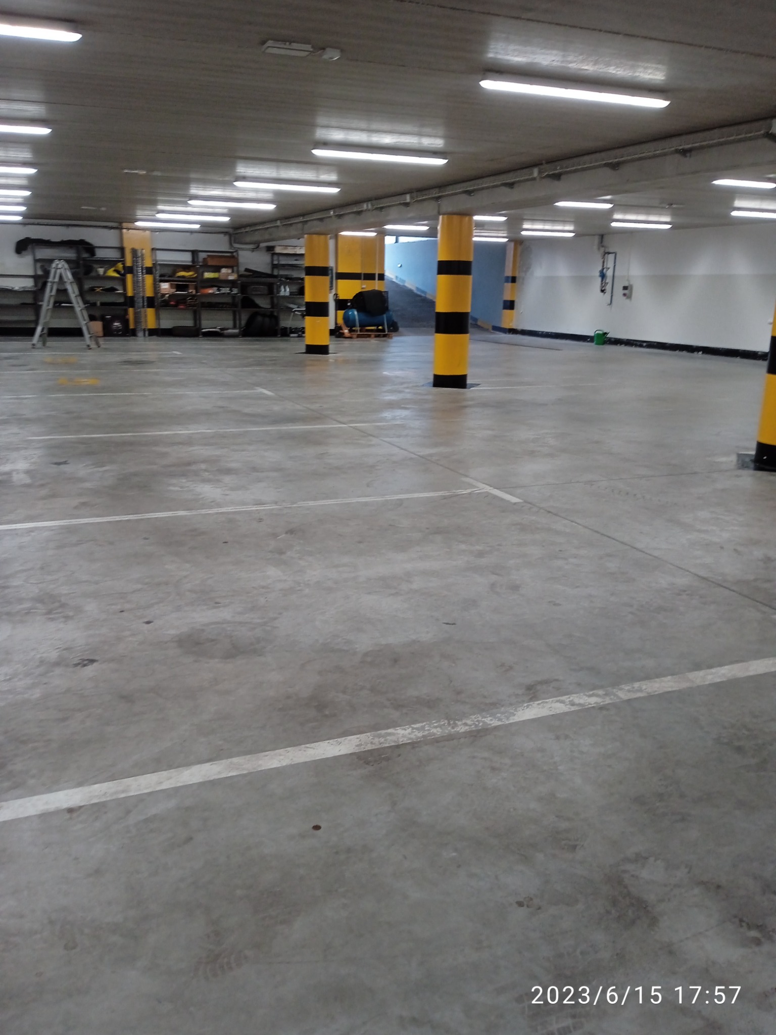 Pusty parking podziemny z betonową podłogą i żółto-czarnymi słupami, oświetlony rzędami lamp fluorescencyjnych, z regałami w tle i osobą pracującą w oddali.