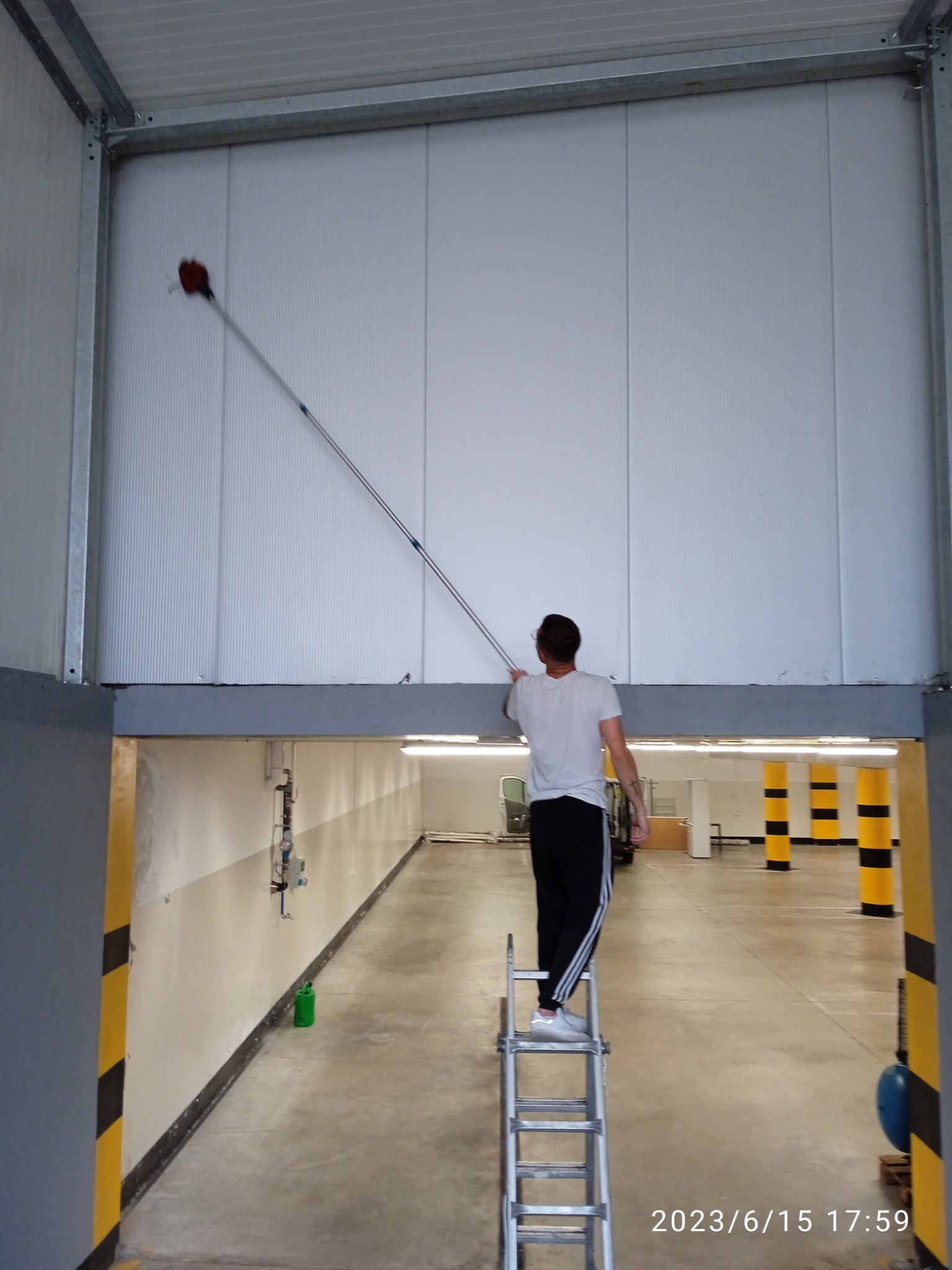 Mężczyzna na drabinie czyści wysoką, białą ścianę w hali za pomocą narzędzia z długim uchwytem i czerwoną końcówką.