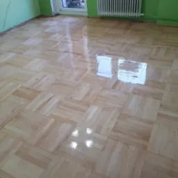 Parkiet ułożony w kwadraty, świeżo polakierowany, z widocznymi refleksami światła, ściany w kolorze zielonym.