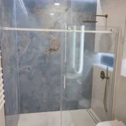 Nowoczesna kabina prysznicowa z przesuwnymi drzwiami, wykończona płytkami imitującymi marmur w odcieniach błękitu i beżu, z elementami armatury w kolorze szczotkowanego złota i podświetleniem LED...