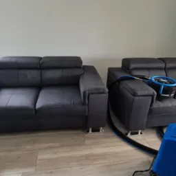 Dwie ciemnoszare kanapy z regulowanymi zagłówkami na metalowych nóżkach, jedna z nich w trakcie czyszczenia za pomocą niebieskiego odkurzacza piorącego z czarnym wężem i okrągłą szczotką na siedzisku.