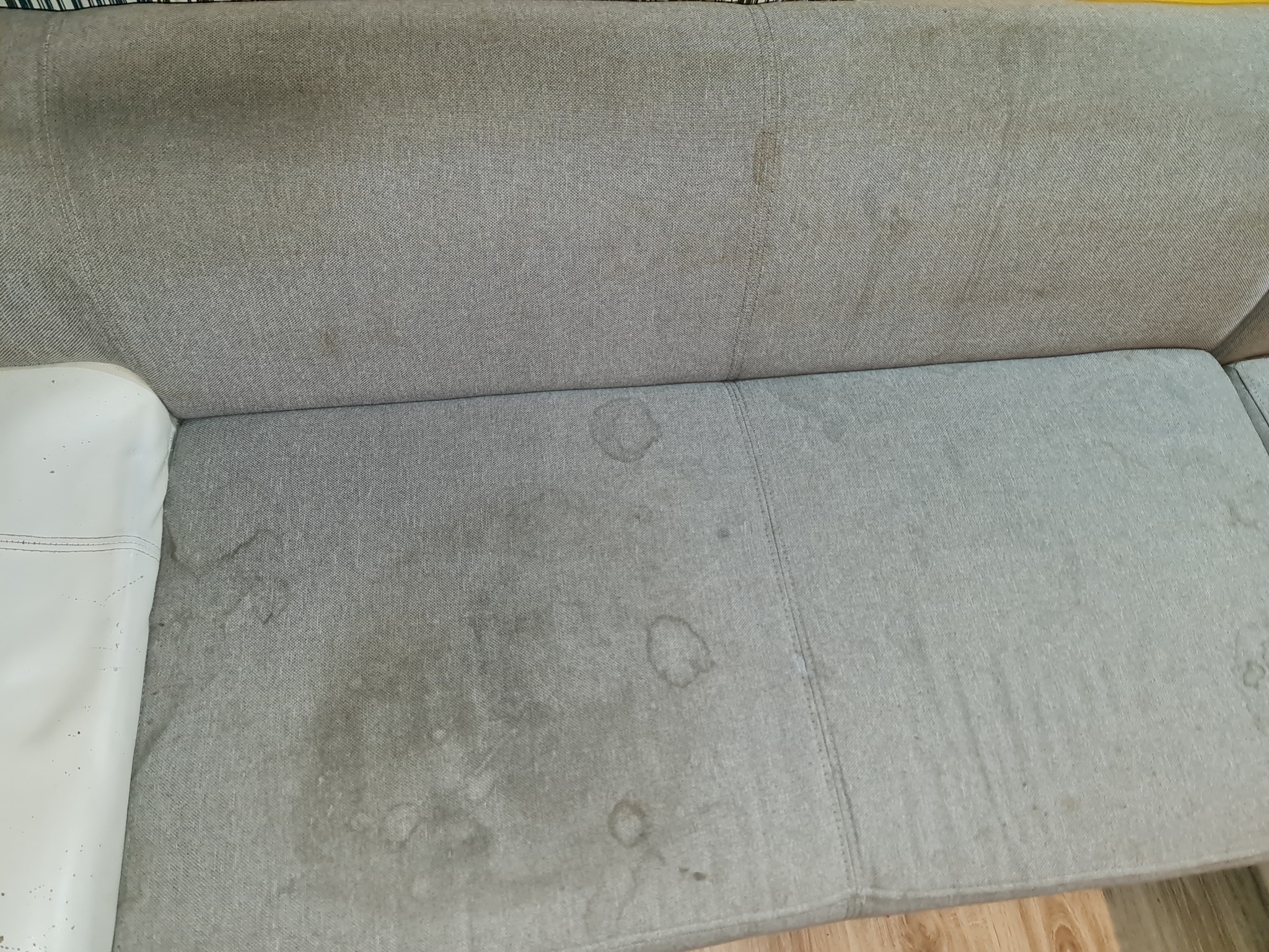 Szara, materiałowa kanapa z widocznymi plamami i zabrudzeniami na siedzisku i oparciu, ukazująca potrzebę czyszczenia tapicerki.