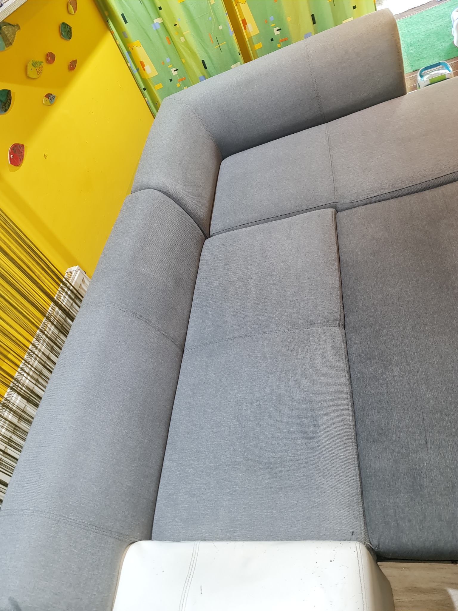 Szara, narożna kanapa z widocznymi plamami na siedzisku, ustawiona w pokoju z żółtą ścianą ozdobioną kolorowymi elementami dekoracyjnymi i zielonymi zasłonami.