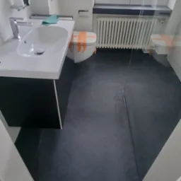 Remontowana łazienka z czarną podłogą z płytek, białą umywalką z szafką, zabezpieczoną taśmą toaletą i grzejnikiem pod oknem z żaluzjami.