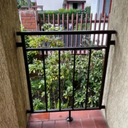 Ciemna, metalowa balustrada balkonowa z widokiem na bujną zieleń ogrodu, widziana z zewnątrz, z mokrą, czerwoną terakotą na podłodze.