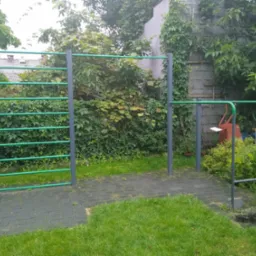 Zewnętrzny zestaw do ćwiczeń z drabinką poziomą i drążkiem do podciągania, malowany na szaro z zielonymi akcentami, umieszczony na trawniku z kostką brukową w tle z zielenią.