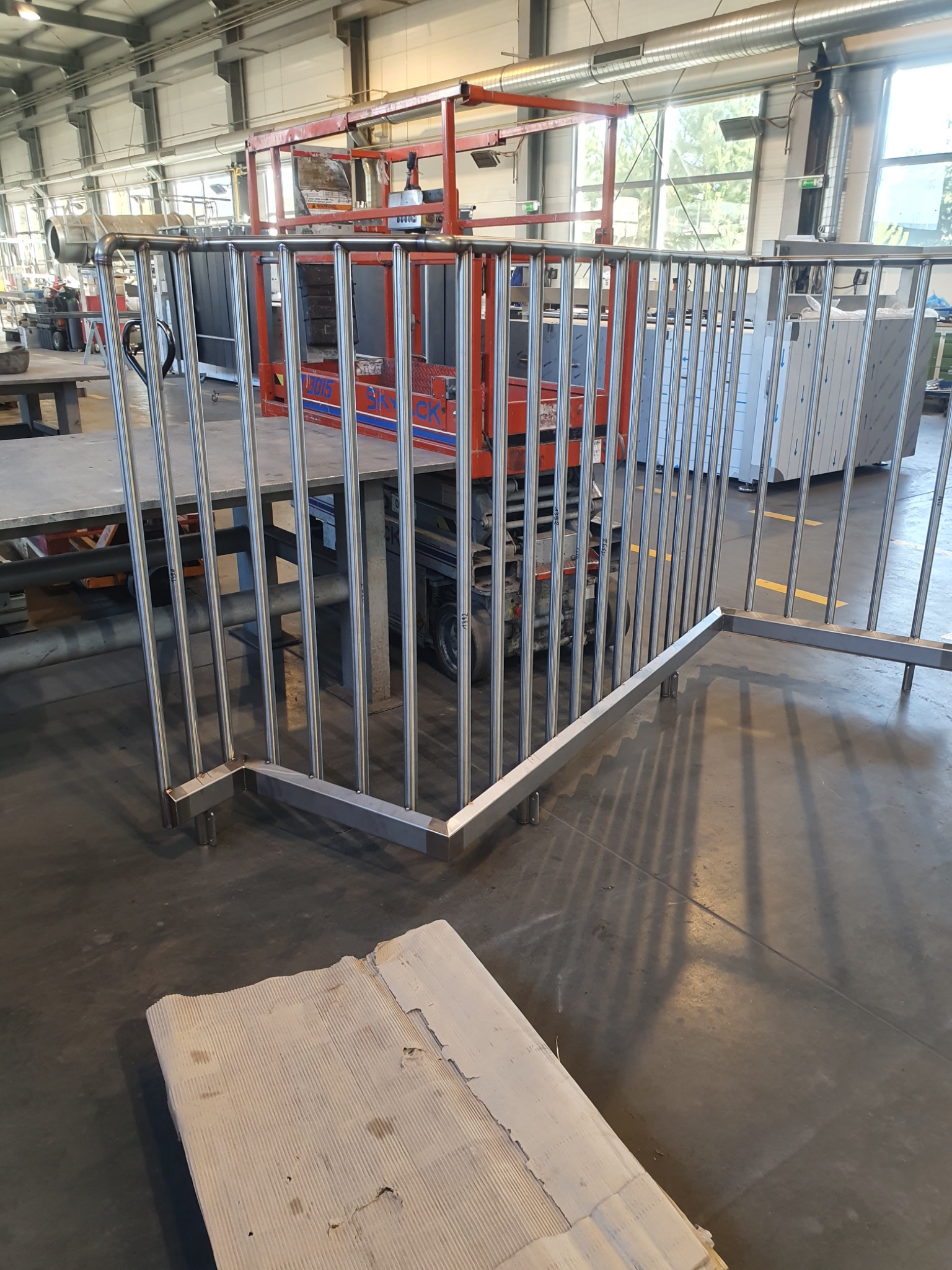 Częściowo zmontowana, surowa, metalowa barierka z pionowych prętów w warsztacie, z widocznym podnośnikiem nożycowym w tle i kartonem na podłodze.