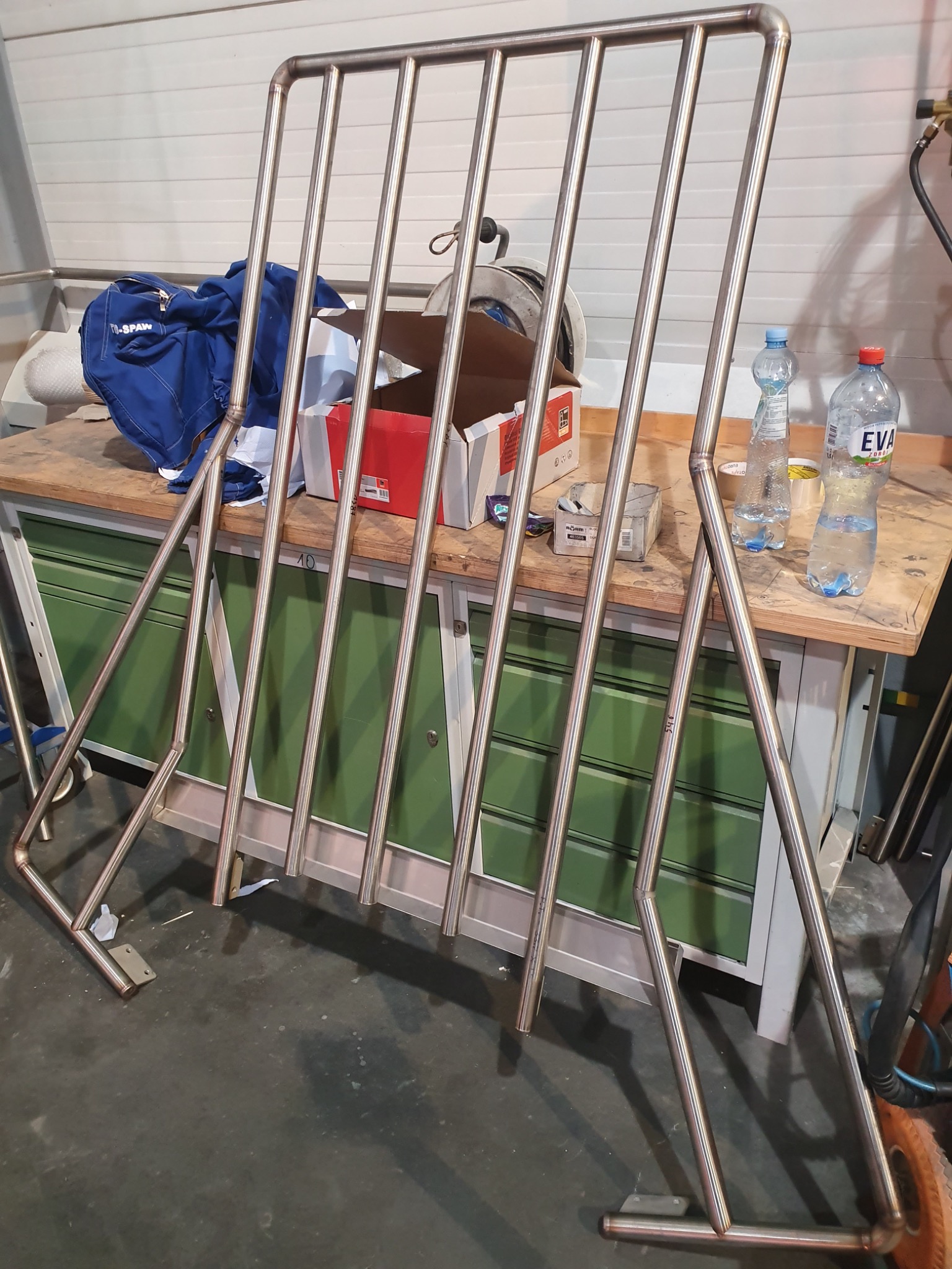 Nierdzewna konstrukcja kratowa z rur, prawdopodobnie balustrada, stojąca na stole warsztatowym w otoczeniu narzędzi i materiałów eksploatacyjnych.