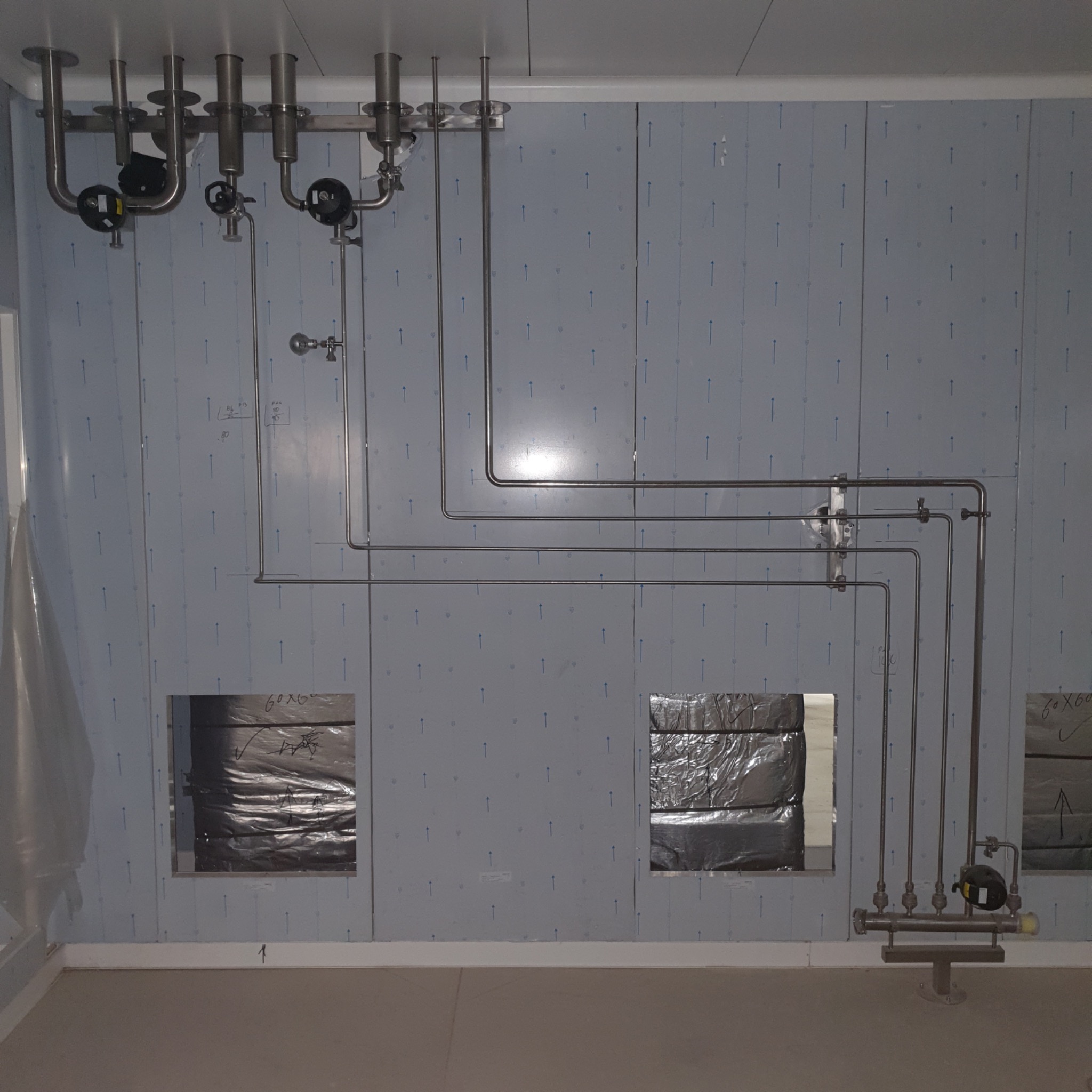 Wnętrze pomieszczenia z zamontowaną instalacją z rur ze stali nierdzewnej, widoczne zawory, liczniki i izolacja w otworach w ścianie.