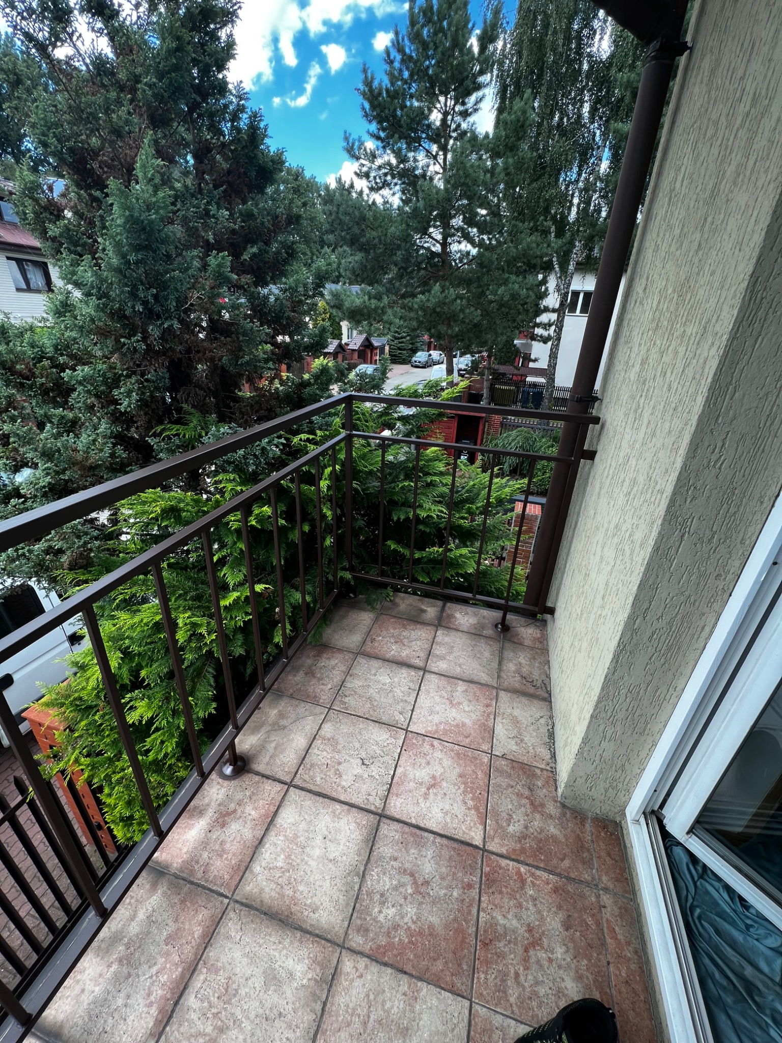 Balkon z brązową metalową balustradą, widok na zieleń drzew i zabudowę jednorodzinną, płytki podłogowe w odcieniach beżu i brązu, fragment okna balkonowego po prawej stronie.